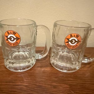 May include: Two clear glass mugs with thick handles and a dimpled base texture. Each mug features a circular orange logo on the front with a black arrow design and the text A&W ROOT BEER printed in black, centered within the orange circle.