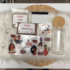 Op de afbeelding: Een assortiment knutselspullen, waaronder glitter, stickers met bemoedigende berichten, een glazen pot met een bamboe deksel, een rakel en twee doorzichtige ringen. De glitter is gelabeld met "Lavish" en "Cinnamon Glaze". De stickers bevatten de woorden "I know my worth" en "Brown Sugar".
