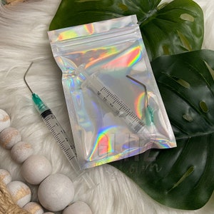 Puede incluir: Una bolsa de plástico transparente con un acabado holográfico iridiscente contiene una jeringa de plástico transparente con una aguja verde y una herramienta de metal curvada con una punta verde.