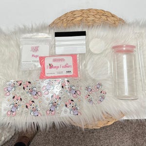 Op de afbeelding: Een heldere glazen beker met een roze deksel, rietje en een Minnie Mouse-sticker. De afbeelding bevat ook een rakel, glitter en roze glitter. De beker is omgeven door andere knutselbenodigdheden.