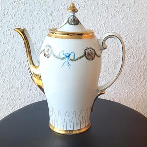 May include: White porcelain coffee pot with gold trim and a blue ribbon design. The pot has a lid with a gold finial and a floral design. The pot is sitting on a black surface.