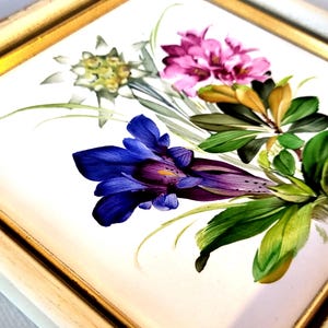 May include: A square ceramic tile featuring a hand-painted floral arrangement with a prominent deep blue gentian flower, pink blossoms, and white edelweiss, all set within a simple gold-toned frame with a white border.