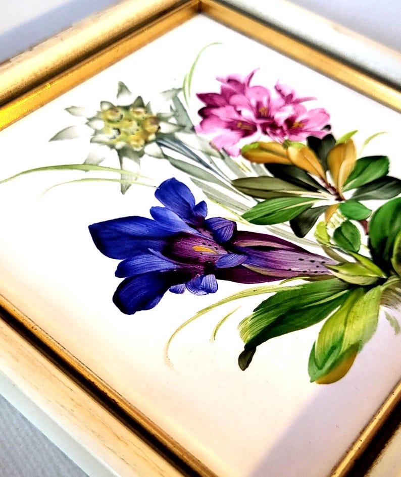 May include: A square ceramic tile featuring a hand-painted floral arrangement with a prominent deep blue gentian flower, pink blossoms, and white edelweiss, all set within a simple gold-toned frame with a white border.