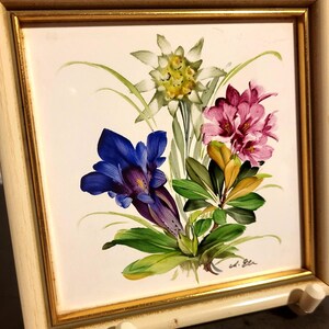May include: A square ceramic tile painting featuring a bouquet of alpine flowers including a white edelweiss, a deep blue gentian, and pink rhododendrons with green leaves. The tile is set in a light-coloured wooden frame with a thin gold inner border.