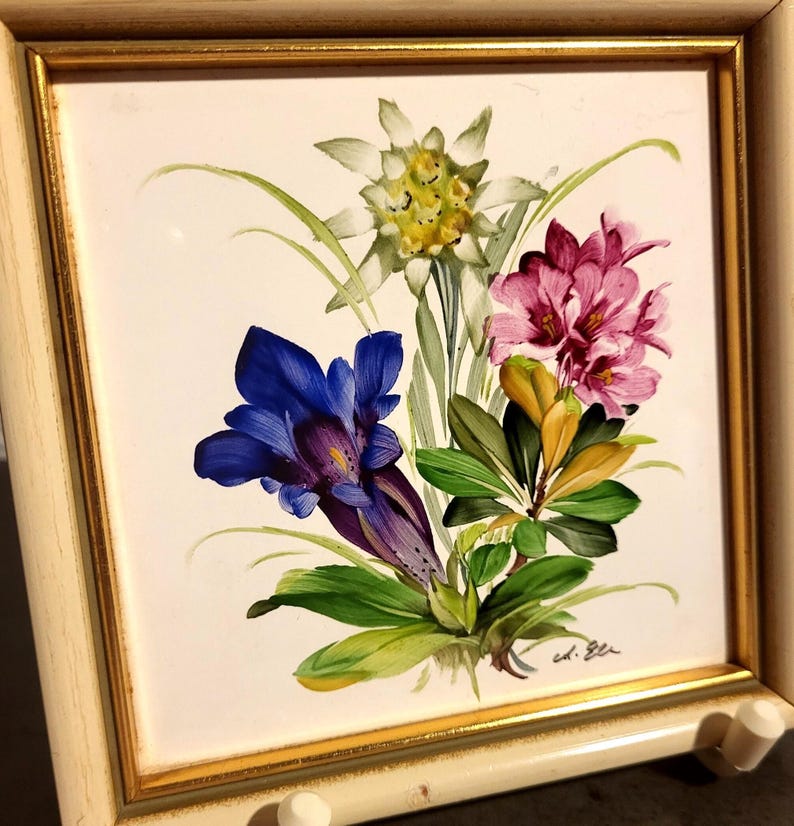 May include: A square ceramic tile painting featuring a bouquet of alpine flowers including a white edelweiss, a deep blue gentian, and pink rhododendrons with green leaves. The tile is set in a light-coloured wooden frame with a thin gold inner border.