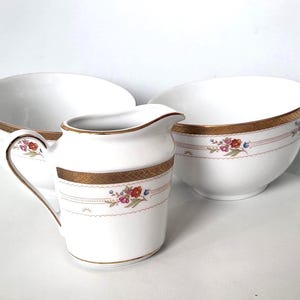 May include: A white ceramic creamer and two bowls. The creamer and bowls have a gold rim and a decorative band with floral and leaf designs in red, blue, and pink. The bowls are different sizes.