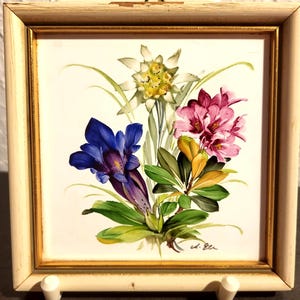 May include: A square wall plaque featuring a painted floral arrangement of white edelweiss, a deep blue gentian, and pink rhododendron blossoms. The artwork is set within a distressed off-white wooden frame with a thin gold inner border.