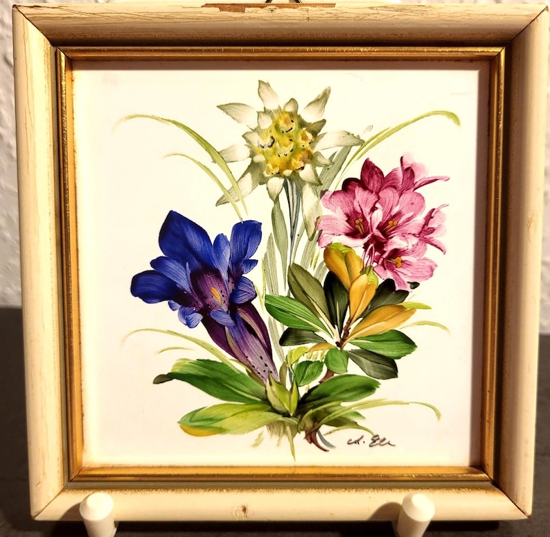 May include: A square wall plaque featuring a painted floral arrangement of white edelweiss, a deep blue gentian, and pink rhododendron blossoms. The artwork is set within a distressed off-white wooden frame with a thin gold inner border.
