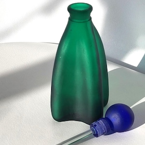 Può includere: Un flacone di profumo in vetro verde con un contagocce in vetro blu abbinato. La bottiglia ha una forma curva unica e una finitura opaca. Il contagocce ha un tubo di vetro trasparente e una lampadina blu rotonda.