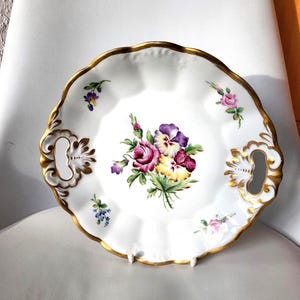 May include: White porcelain serving plate with a gold rim and handles. The plate features a floral bouquet of purple, pink, and yellow flowers. Small floral accents are scattered around the edge of the plate.