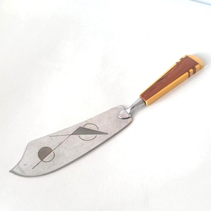 May include: A vintage cheese knife with a stainless steel blade and a decorative handle. The handle features light yellow and dark brown wood. The blade has a geometric design.