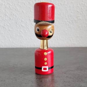 Könnte beinhalten: Eine Holzfigur eines Soldaten mit roter Mütze und Uniform. Die Figur hat ein braunes Gesicht mit roter Nase, schwarzem Schnurrbart und geschlossenem Auge. Die Uniform hat goldene Knöpfe und eine weiße Gürtelschnalle.