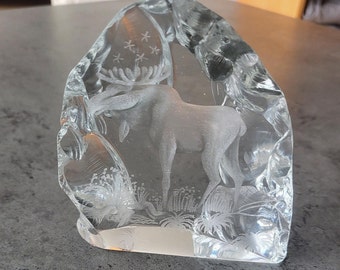May include: A clear glass paperweight with an etched design of a moose standing in a forest. The moose is facing the left side of the image and has a large set of antlers. The forest is filled with trees and plants. There are five stars etched in the top of the paperweight.