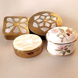 May include: A collection of four small trinket boxes: a square brass box with mother-of-pearl inlay, a round brass box with geometric mother-of-pearl inlay, a round gold-tone metal box with a mother-of-pearl lid, and a white porcelain box with floral motifs.