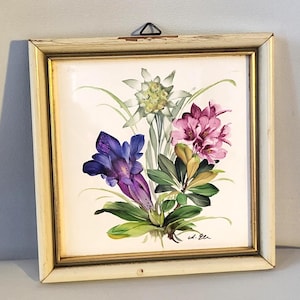 May include: A square wall plaque featuring a hand-painted floral arrangement of a white edelweiss, a deep purple gentian, and pink rhododendron blossoms. The artwork is encased in a distressed off-white wooden frame with gold trim.