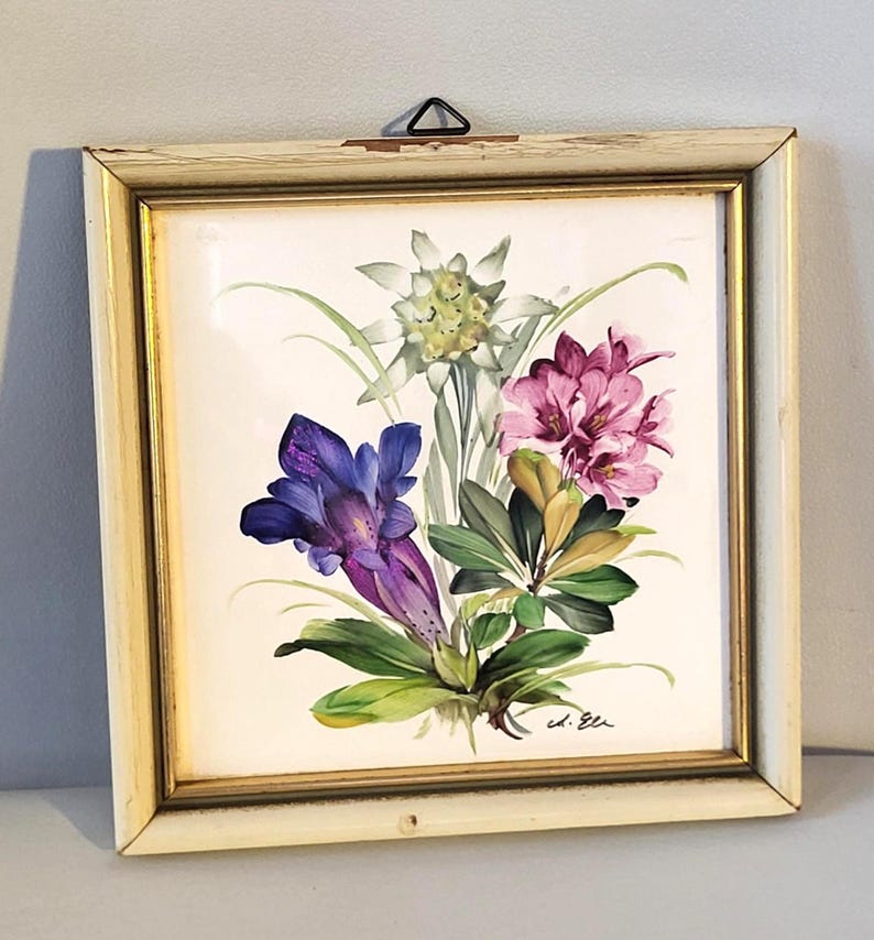 May include: A square wall plaque featuring a hand-painted floral arrangement of a white edelweiss, a deep purple gentian, and pink rhododendron blossoms. The artwork is encased in a distressed off-white wooden frame with gold trim.