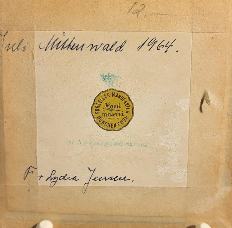 May include: A square beige paper label featuring a yellow circular sticker with the text Porzellan-Manufaktur M&uuml;nchen GmbH Handmalerei. Handwritten text includes Juli Mittenwald 1964, the number 12, and the name F. Lydia Jensen.