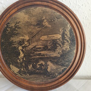 May include: A framed print depicting a hunting scene with a man firing a gun at a bird in flight. The print is in black and white and has a gold background. The frame is made of dark wood.