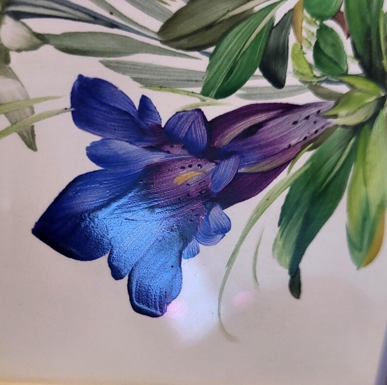 May include: A hand-painted illustration on a white surface featuring a vibrant blue and purple bell-shaped flower with visible brushstrokes. The flower is surrounded by long, pointed green leaves and stems, creating a detailed botanical composition.