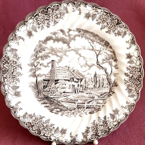 Puede incluir: Un plato de cena de cerámica blanca con un patrón floral marrón y blanco alrededor del borde. El centro del plato presenta una ilustración en blanco y negro de una cabaña en un entorno rural.