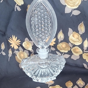 May include: A clear, vintage perfume bottle with a decorative, leaf-shaped stopper. The bottle has a detailed, textured design and a fan-shaped base. The stopper is oval with a diamond pattern. The bottle is made of glass.