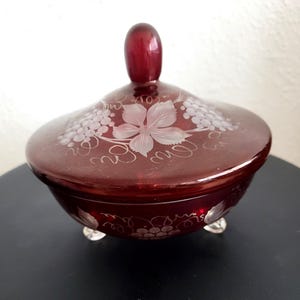 Puede incluir: Un plat de dulces de vidrio rojo con tapa, decorado con un diseño de vid grabado. El plato tiene tres patas y una perilla en la tapa.