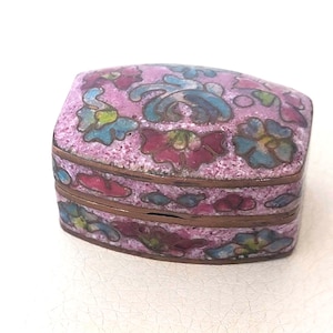 May include: A small, oval-shaped metal trinket box featuring a pink enamel base with a multi-colored floral pattern in shades of blue, red, and green. The box has a hinged lid and a metallic rim that separates the lid from the base section.