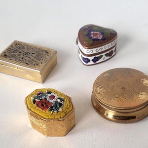 May include: Four antique decorative boxes. A rectangular gold box with an engraved design, a heart-shaped box with floral enamel, a mosaic box with a floral design, and a round gold box with a floral pattern.