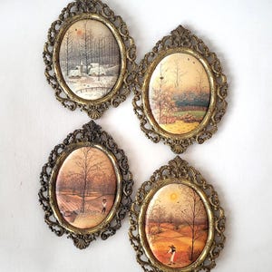 May include: Four oval wall plaques featuring landscape scenes in ornate, metallic gold-toned frames. Each plaque depicts a different seasonal view, including a snowy forest, a green field, a barren tree landscape, and a sunlit autumn meadow.