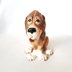 May include: A ceramic Basset Hound dog figurine, predominantly brown and tan, with a black nose and a mournful expression. The dog is depicted in a seated pose, with cream-coloured paws and chest. A decorative item, suitable for display.