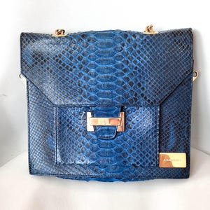 May include: A rectangular shoulder bag featuring a blue and black snakeskin-textured pattern. The bag includes a gold-toned metal clasp on the front flap, a gold-toned rectangular plaque in the bottom right corner, and gold-toned chain shoulder straps.