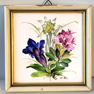 May include: A square wall tile featuring a hand-painted floral arrangement of a white edelweiss, a deep blue gentian, and pink rhododendron blossoms with green leaves, set within a distressed off-white wooden frame with gold trim.