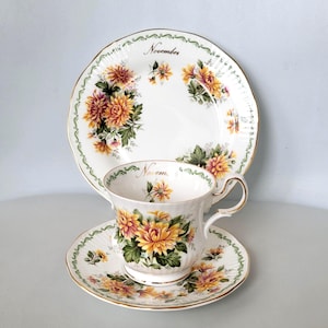 May include: A white teacup, saucer, and plate set with a floral design featuring yellow and orange chrysanthemums. The set is decorated with a green and gold trim and the word "November" is written on the plate.