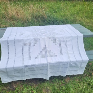 以下が含まれることがあります： 幾何学模様の繊細な刺繍が施された白いテーブルクロス。テーブルクロスは木製のテーブルの上に置かれています。