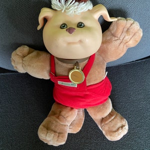Peut inclure: Un jouet en peluche en forme de chien brun et blanc, portant une tenue rouge et blanche. Le chien a un nez brun, des yeux bruns et un collier blanc avec une étiquette dorée qui dit "Love you lots".