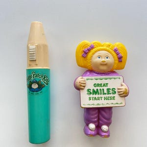 May include: Two vintage Cabbage Patch Kids collectibles. One is a teal and beige crayon-shaped item with the Cabbage Patch Kids logo. The other is a doll figure holding a sign that reads "Great Smiles Start Here".