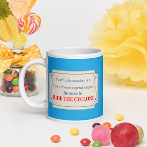 May include: A white ceramic mug with a bright blue background featuring a rectangular label with the text: Your lucky number is 7. You will soar to great heights. Be sure to RIDE THE CYCLONE. The mug is surrounded by colourful sweets and a yellow pom-pom.