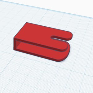 Puede incluir: Un objeto rojo en forma de U con una base rectangular y bordes redondeados. El objeto parece ser un diseño impreso en 3D, posiblemente un soporte o un clip. El fondo es un patrón de cuadrícula.