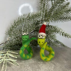 Könnte beinhalten: Zwei Filz-Schmuckschlangen. Eine ist einfarbig grün, die andere limettengrün mit dunkleren grünen Punkten und einer roten Weihnachtsmütze. Beide haben Wackelaugen und hängen an weißen Schnüren, ideal für die Weihnachtsdekoration.