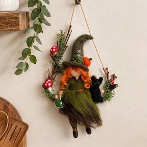 May include: A needle-felted witch doll with bright orange hair and a green dress, wearing a pointed hat. The doll is suspended on a twig wreath decorated with green moss, red-spotted mushrooms, and a small black cat, holding a tiny green potion bottle.