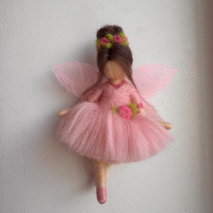 May include: A felt fairy doll in a pink dress and wings, with brown hair and a floral crown. The doll is handmade and suspended against a white background, showcasing a whimsical design.