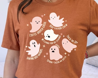 May include: A rust-colored t-shirt featuring a circular design of cartoon ghosts and text. The ghosts are in various poses and colors, with phrases like "Coffee Driven Ghoul" and "Spook-tacular Data". The design is surrounded by small stars.