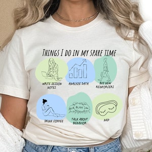 Puede incluir: Camiseta color crema con el texto "THINGS I DO IN MY SPARE TIME" e ilustraciones de actividades como escribir notas, analizar datos y tomar una siesta. La camiseta tiene cuello redondo y mangas cortas.
