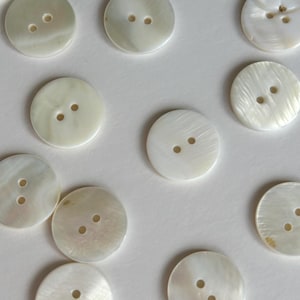 Può includere: Una collezione di bottoni rotondi bianco sporco con due o quattro fori. I bottoni hanno una lucentezza madreperlacea e sono disposti su una superficie bianca. Adatti per cucito e progetti di artigianato.