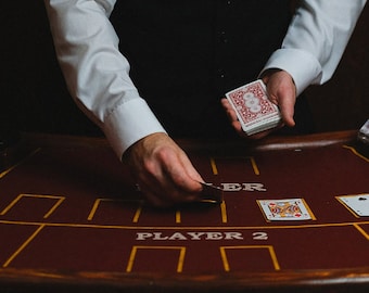 May include: A person wearing a white shirt and a black vest is dealing cards at a red felt gaming table. The table has a white inscription that reads "PLAYER 2".