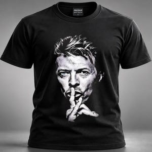Op de afbeelding: Een zwart T-shirt met korte mouwen en een grote, contrastrijke zwart-wit fotoprint van een persoon met één blauw oog, die een vinger voor de lippen houdt in een stiltegebaar. Het shirt heeft een ronde hals en een Gildan-merklabel.