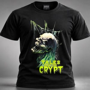 Peut inclure: Un t-shirt noir à manches courtes avec un motif représentant une créature squelettique pâle aux yeux brillants et aux cheveux verts. Au-dessus de la créature se trouvent des lignes fines en forme de toile. Le texte TALES FROM THE CRYPT est en vert.