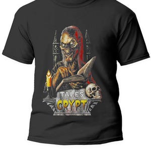 Peut inclure: T-shirt noir avec un graphique d'une figure effrayante tenant un livre. La figure est entourée d'une arche de pierre et le texte "Tales from the Crypt" est sous la figure.