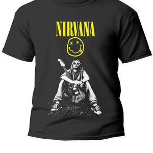 Peut inclure: T-shirt noir avec le logo Nirvana jaune et un graphisme d'une personne avec une guitare. La personne est assise, tenant la guitare. Le t-shirt est de style col rond.
