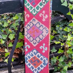 May include: A tall, cylindrical carrying case featuring a vibrant red woven fabric with a repeating geometric pattern in blue, green, and orange. The bag includes a black adjustable shoulder strap and a drawstring closure with red and blue beads.